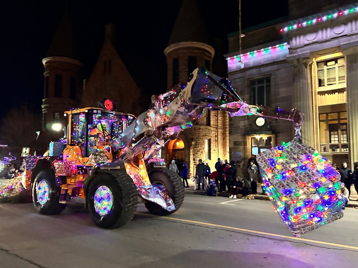 Victoria Street at night lined with spectators and a piece of heavy equipment drives by adorned in illuminated christmas lights